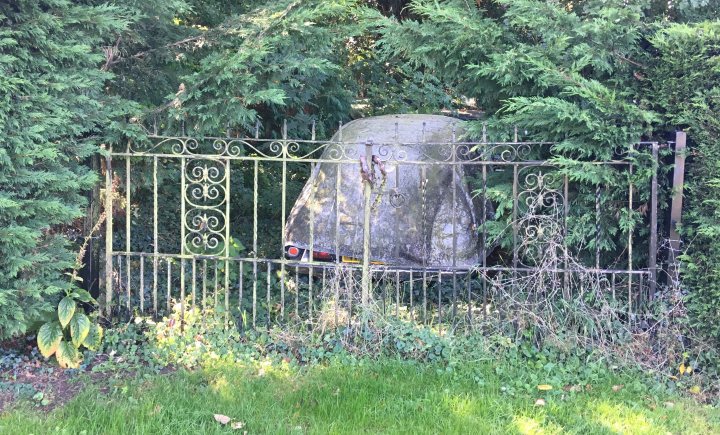 A wooden bench sitting in the middle of a forest - The image shows a wrought iron gate in the foreground with a rustic stone object placed behind it. This stone object resembles a small wall or structure that has an old, weathered appearance. Behind the gate and object, there is a lush green garden filled with various plants and trees, suggesting a well-maintained outdoor space. The background features more greenery, including leafy trees, indicating that this scene is likely set in a rural or suburban area during what appears to be late spring or early summer, given the full bloom of the plants.