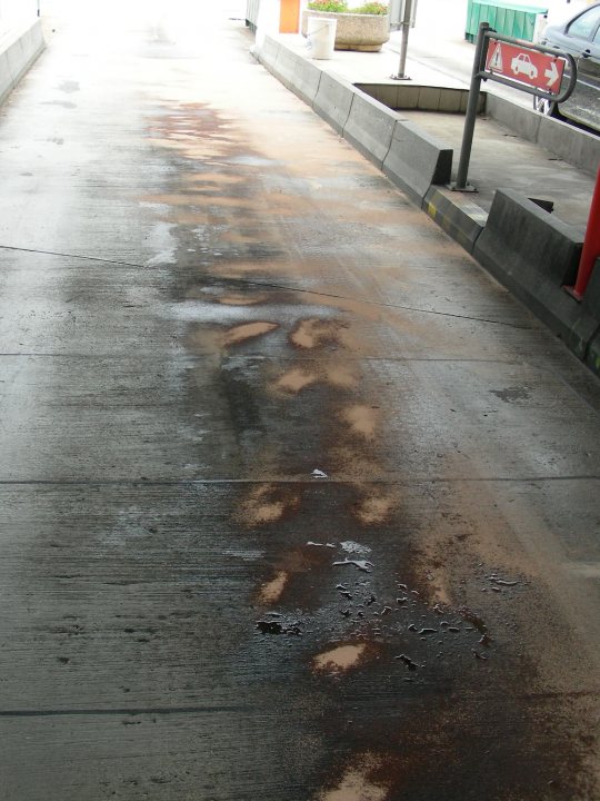 Can anyone help? - Page 1 - France - PistonHeads - The image shows a concrete surface with numerous tire tracks and patches of dark puddles. Part of a road or plaza, it appears to be a sunny day given the shadows and the way the concrete soaks up the sunlight. There's a red roadblock visible on the right side of the image, indicating a possible traffic restriction or safety measure. In the background, there's a parking meter, suggesting this area might have regulated parking.