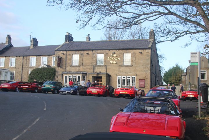 A car parked on the side of the road - Pistonheads - The image shows a street scene with a two-story brick building that appears to be named "The Crown Hotel." There are numerous cars parked along the street, predominantly parked facing opposite directions, indicating a one-way street with parking allowed on both sides. The cars at the forefront are a mix of red and silver, and there are no people visible. The sky is blue and poles with signs are seen near the curbs. The overall impression is of a calm, possibly rural or suburban, setting.