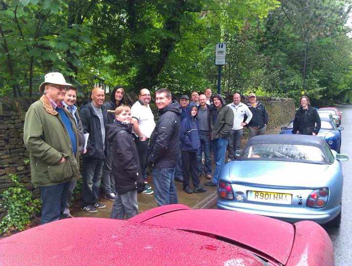 Peak District Run Sunday May 25th - Page 3 - TVR Events & Meetings - PistonHeads - The image portrays a lively gathering of people standing outside. A diverse group of adults, including both men and women, are clustered around the scene. They appear to be grouped near parked cars, suggesting a casual, outdoor setting. A few individuals are under umbrellas, perhaps indicating recent rain. There's a cake spotted among the group, hinting at a celebration or special occasion. Overall, the image captures a moment of shared experience among the group.