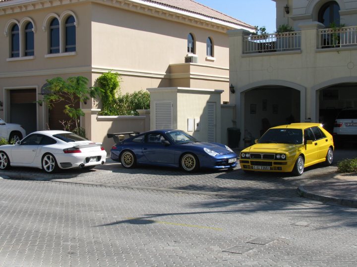 Drives Readers Pistonheads Middle East - The image shows a serene urban scene, highlighting a row of parked cars on a gray brick street. On the left, there's a majestic white mansion with towering columns, its stark simplicity contrasting with the vibrant green foliage on the front lawn. The middle of the row is occupied by a striking blue car, its sleek design amplified by the chrome rims. To the right, a sunshine yellow sports car stands out against its surroundings. The sky above is a peaceful blue, completing this picturesque setting.