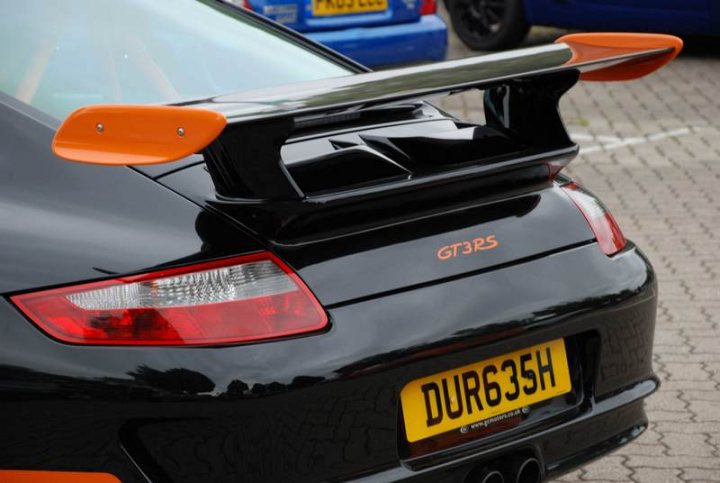 Pistonheads Service Sunday - The image showcases a sleek black sports car parked in a gravel-covered lot. The car's license plate reads "DUR635H." Its design features extended wings, a sporty rear spoiler, and a dual exhaust pipe. In the background, there are other vehicles, including a blue car and another that's not clearly visible.