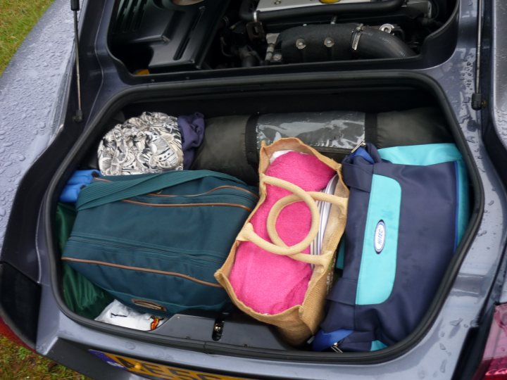 Pics of your bootspace pls - Page 1 - Elise/Exige/Europa/340R - PistonHeads - The image features an open trunk of a car filled with various items. Some of the items include a blue bag, a pink mat, a black backpack, and a reusable shopping bag with a white stripe. The trunk appears to be wet, suggesting recent rainfall. The overall scene conveys a sense of travel or outdoor activity preparation.