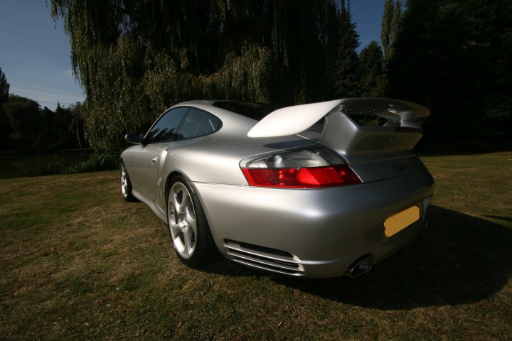 Words Worth Pistonheads - The image shows a silver Porsche with a distinctive rear-end design, parked in what appears to be a park or wooded area during the day. The car's bodywork is gleaming, with an open rear tailgate showing various air vents and what looks like a plaid design on the seat. The background features a serene setting with natural scenery, including a grassy area and some trees, contributing to a peaceful and quiet atmosphere.