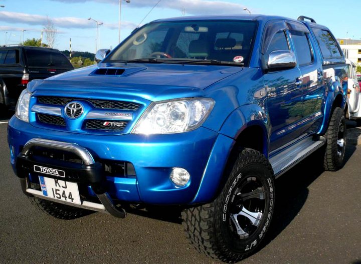 Pistonheads - The image showcases a stylish blue Toyota Land Cruiser parked on a road. The vehicle is positioned at an angle to the camera, providing a side view. The Land Cruiser is equipped with large off-road tires and a roof rack, signifying its robust build and capabilities. The license plate clearly displays numerical information. The overall vibrant blue color of the truck contrasts well against the outdoor setting, and the truck appears to be in excellent condition.