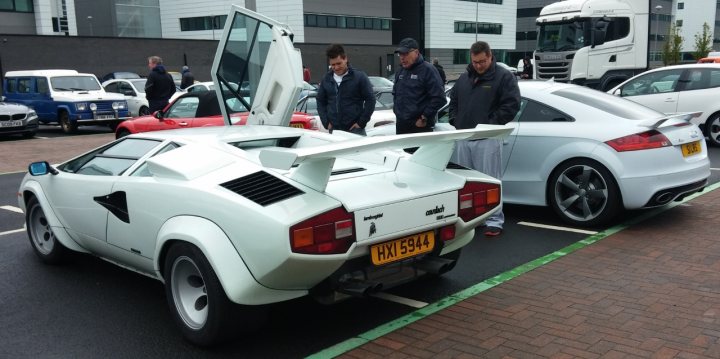 A police car is parked on the side of the road - Pistonheads - In the image, a group of men is gathered in a parking lot, admiring several cars. One of the cars stands out due to its unique feature: a large white spoiler attached to its back. The men appear interested in this particular car, possibly discussing its design or performance, as they are standing next to it. The environment suggests a casual and friendly atmosphere. The clothing of the men varies, indicating a relaxed, informal gathering. The overall scene conveys a shared interest in automobiles.