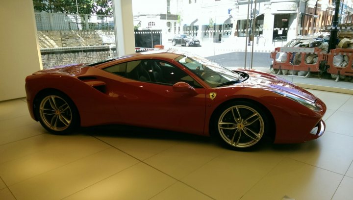 Only UK 488 GTB... - Page 2 - Ferrari V8 - PistonHeads - The image depicts a bold and vibrant red sports car, possibly a Ferrari, stationed on a reflective tiled floor inside a spacious garage or showroom. The vehicle is sleek and has minimal detailing, with two doors and a sloping roofline. The headlights and taillights are discreetly integrated into the body of the car, highlighting its aerodynamic and sporty design.

The showroom itself appears to be well-lit, with large windows that offer a view of the outdoors where cars are parked. The indoor environment contrasts with the cars parked outside, emphasizing the car's presence in the showroom. The car's expertly crafted details suggest it is a luxury vehicle, adding to the overall impression that the image is a representation of luxury and exclusivity.