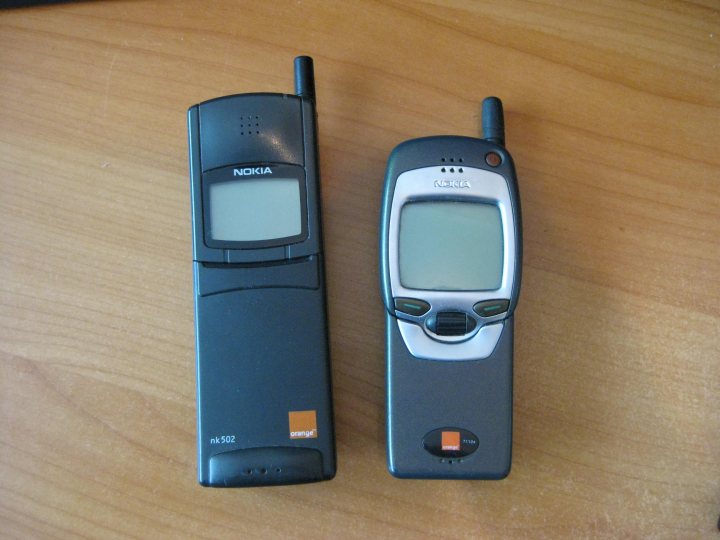 Phone Banana Content Ringringringringring Geek Pistonheads - The image shows two open flip cell phones lying next to each other on a wooden surface. The phone on the left is traditional black and gray while the one on the right has a modern gray and black color scheme. Both phones have distinctive hinges and external screens. The phones are depicted lying flat with the hinges facing upwards and the screens facing downwards. There are visible brand details and a charging port on the lower part of both phones. The image is simple and straightforward, focusing solely on the items without any additional context or background.