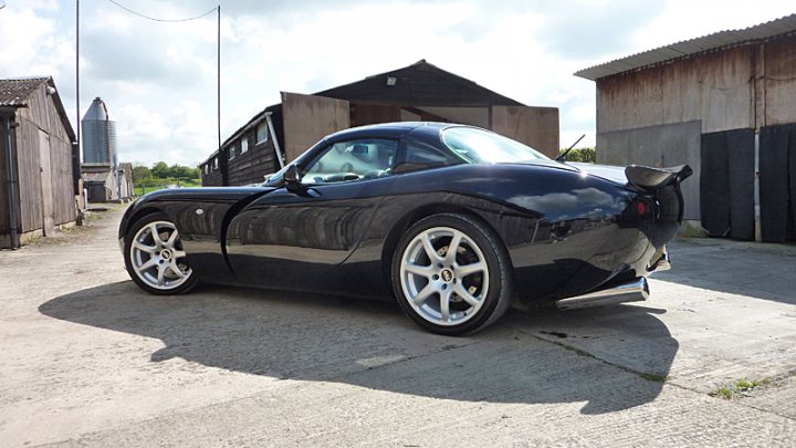 WHICH CLASSIC 4X4 FOR UP TO £6K - Page 1 - Off Road - PistonHeads - The image shows a sleek, dark-colored sports car parked on a concrete surface, which is lined with small structures that resemble sheds. The car is positioned slightly to the left of the center line, giving a view of its right side. It has a low, accentuating the aerodynamic design typical of high-performance vehicles. The sky appears overcast, suggesting a cloudy day, and the environment suggests a rural or semi-rural setting due to the presence of the sheds.