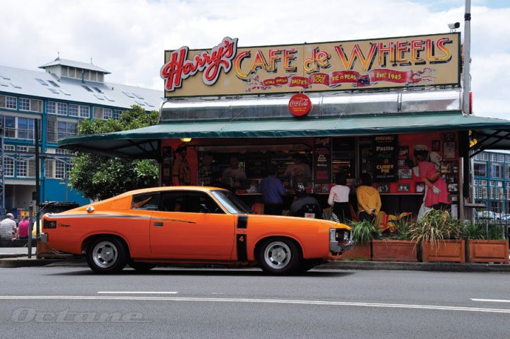 Pistonheads Americanaustralian - This image captures a street scene featuring a small but distinctive orange car parked in front of a local business. The business, presumably a cafe, has a sign reading "Harry's Cafe de Wheels," indicating it might be a diner or a place where food is served in a cozy setting. The atmosphere appears casual, with a few people leisurely strolling by, and the car is parked on the side of the road, waiting patiently for its owner to return. The overall image gives a sense of everyday life in this part of town.