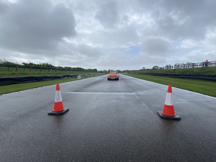 Pictures of your Classic in Action - Page 23 - Classic Cars and Yesterday's Heroes - PistonHeads UK - This is a color photograph showing an empty road with two orange traffic cones. The cones are situated in the center of the frame, with a black and white checkered pattern beneath them on the asphalt. The surrounding area appears to be wet due to recent rainfall, and there are no visible vehicles or pedestrians. In the background, we can see a landscape with greenery under an overcast sky. There is also a fence running along the left side of the road, suggesting that this might be a designated track or closed area for driving.