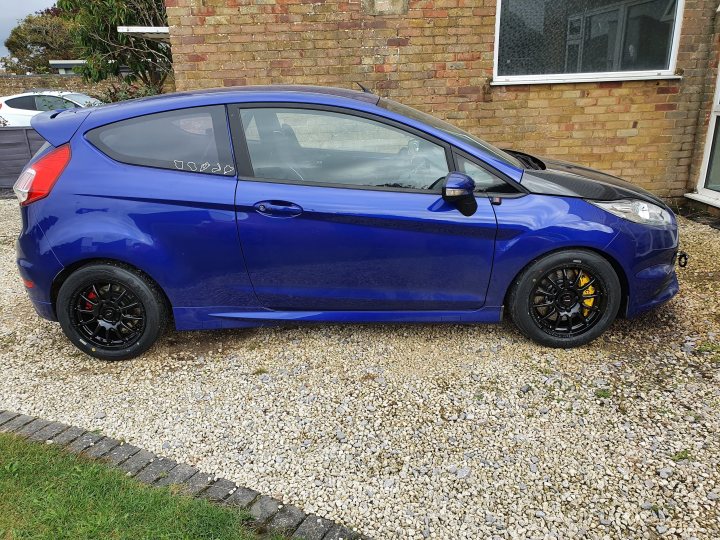 Fiesta ST MK7.5 Track car  - Page 31 - Readers' Cars - PistonHeads UK - The image features a bright blue Ford Fiesta parked on a gravel driveway. The car is equipped with a black spoiler and black rims, giving it a sporty appearance. It appears to be well-maintained, with no visible damage or scratches. In the background, there's a building with a white exterior and a visible window. The vehicle has a license plate, but the text is not legible in this image.