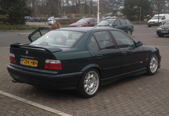 My 1997 E36 328i - Page 1 - Readers' Cars - PistonHeads - The image shows a dark-colored car parked on what appears to be a brick surface. The car is a hatchback with a visible license plate and a pair of side mirrors. A person can be seen in the background, and there are various other cars parked or moving nearby, indicating this might be a parking lot or a busy street. The car is the central focus of the image.