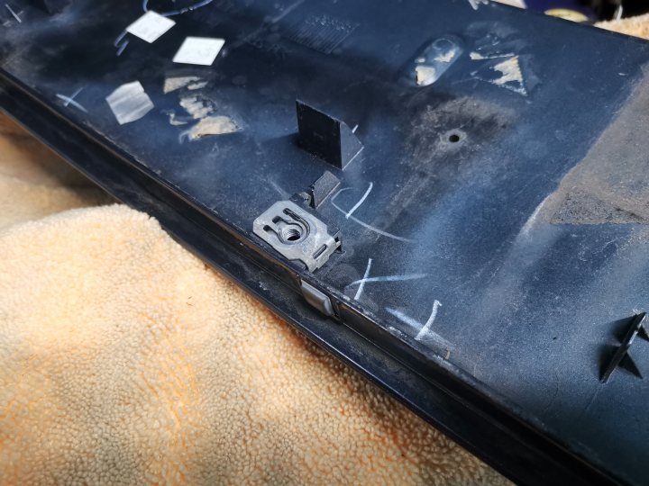 The image shows a damaged black smartphone with a cracked back screen. It is lying on its side, displaying the damaged casing and the internal circuitry beneath it. The phone appears to be discarded, as indicated by a pile of dust on top. The background suggests an indoor setting, possibly a workshop or a repair shop, given the presence of tools.