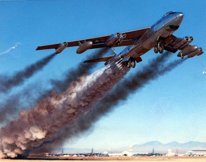 Pistonheads Aircraft Amazingly - The image displays a dramatic scene of a large airplane hovering near the ground, emitting a significant amount of smoke that billows into the sky. The airplane appears to be a military transport aircraft, possibly a jumbo jet, due to its size and distinctive markings on the wings. The sky is a clear, blue expanse, providing a stark contrast to the black smoke. In the distance, several mountains can be seen, adding a sense of scale and location to the photograph. The image captures a moment of intense activity, likely indicating the aircraft's takeoff process with a mixture of air pollutants.