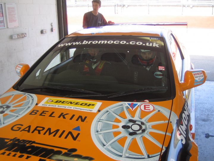 Btcc Pistonheads Bromocohalfords - The image features a unique orange car, prominently emblazoned with various logos including "www.bromco.co.uk," "Belkin," "Garmin," and "Simmons." The car appears to be in a garage or workshop, with men seated inside looking out at the camera. The style of the image seems candid, possibly taken during a break or a casual moment in the day of these individuals. The car, contrasting in color with the indoor setting, is the central focus against the darker background, suggesting it's a vehicle of interest or importance.