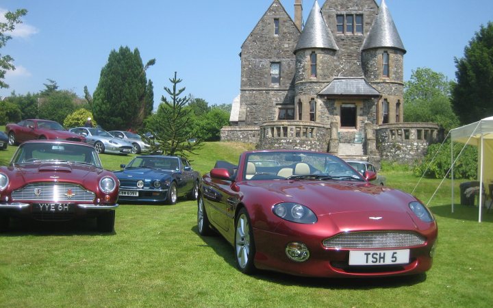 Aston Martin owners Pistonheads Roll call - Page 8 - Aston Martin - PistonHeads - The image presents a stunning scene of a castle, displayed against a backdrop of a clear blue sky. In the foreground, a vintage convertible sports car, specifically a Jaguar, is parked prominently on the grass. The car exudes an aura of nostalgia, complementing the historical charm of the castle. In the distance, there are other classic cars parked around the castle, adding to the vintage atmosphere. The castle itself appears ornate and tall, with a pointed roof characteristic of medieval architecture. The cars and the castle, while distinct, blend harmoniously in the image, creating a picturesque setting.