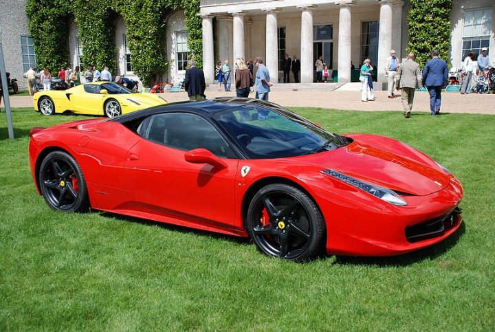 GRRC Lunch Club - Page 1 - Goodwood Events - PistonHeads - The image captures a scene on a well-manicured grass lawn. Two luxury sports cars, one red and the other yellow, are parked side by side. The red car is in the foreground and the yellow car is partially visible behind it. They are surrounded by people, some closer and others a bit distanced, walking on the grass and possibly admiring the cars. The cars are positioned between two white buildings, suggesting this could be a large event venue or a luxury resort. The event appears to be outdoors, with ample space for the participants and the vehicles.