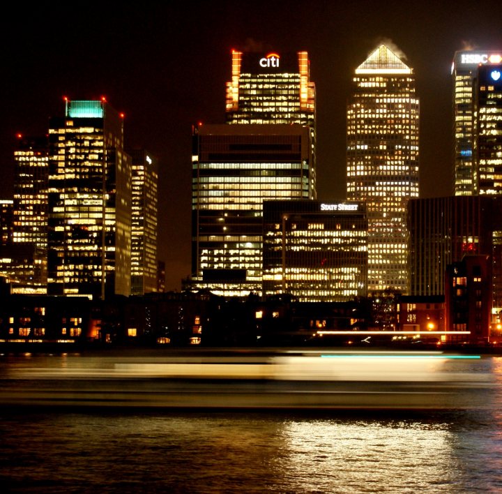 A view of a city street at night - Pistonheads - The image captures a nighttime cityscape with a line of skyscrapers dominating the skyline. The buildings are illuminated, casting light onto the city street below. The city is bustling as evidenced by the blur of a moving vehicle on the top right side of the image, adding a dynamic element to the otherwise static architecture. The photo appears to be taken from across a large body of water, reflecting the lights of the city and providing an extra layer of depth to the scene.