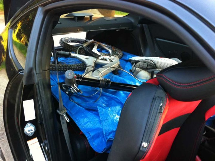 RE: Subaru BRZ RA Racing revealed - Page 4 - General Gassing - PistonHeads - The image shows the interior of a car dealing with a cluttered and cramped space. We can see various items in the trunk, including a bicycle snugly packed alongside a spare tire. Amidst the disarray, a red seat is visible with a secure grin on it. An apartment parking lot is faintly visible in the background with trees peeking into the image. The scene conveys the challenge of organizing and accessing items in the trunk of the car.