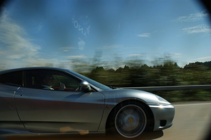 360 thread - Page 1 - Ferrari V8 - PistonHeads - The image captures a silver car, seemingly a speeding Ferrari, as suggested by its curved shape and the luxurious detailing. The car features sleek tires and a tinted window, with a driver visible near the steering wheel, wearing sunglasses. The vehicle is on a road with a clear blue sky overhead and trees lining the street. Populated by several clouds in the sky, the image gives the impression of a fast, possibly thrilling, drive on an open highway. The perspective appears to be from inside the car, as though viewing the scene from the passengers' viewpoint.