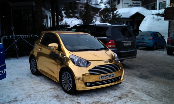 Pistonheads - The image shows a striking gold-colored car parked on the side of a snow-lined road. The car is compact in size and features modern styling, giving it a sleek and luxurious appearance. It is parked in front of a vehicle with a ski rack on top, suggesting it may be used for winter sports. In the background, there are other parked vehicles, including a blue car and another vehicle with a ski rack, which are partially covered with snow due to the winter conditions. The buildings in the background have a charming architectural style, though the winter conditions make it difficult to appreciate the details.