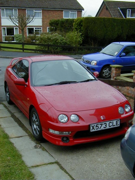 What's your favourite Jap classic ?  - Page 4 - General Gassing - PistonHeads - The image shows a vibrant red sports car parked on a street next to a blue two-door sedan. The car has a compact design with a sleek front and black wheels. It appears to be a sunny day, and the car is facing us, showcasing a european license plate. Behind the red car, there is a sidewalk leading to a parked blue car and a house.