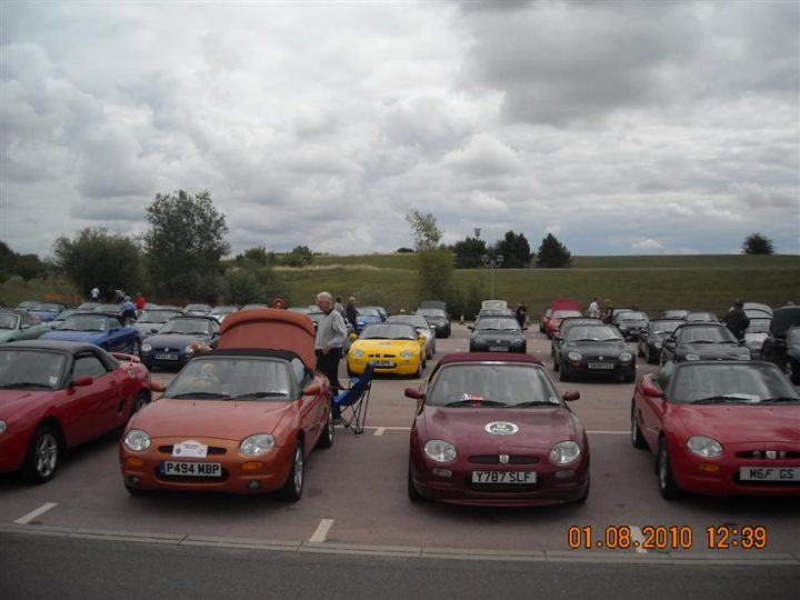 Pistonheads - The image captures a parking lot filled with various cars, each with a distinct color. The most striking are the two red sports cars, one convertible and the other a hard-top, each parked with the top down, inviting other drivers to admire their beauty. A few other cars are also present, parked at different angles, adding to the chorus of colors and models. In the background, the lush greenery of trees and grass contrasts with the concrete lot and creates a serene backdrop for the parcade of impressive vehicles.