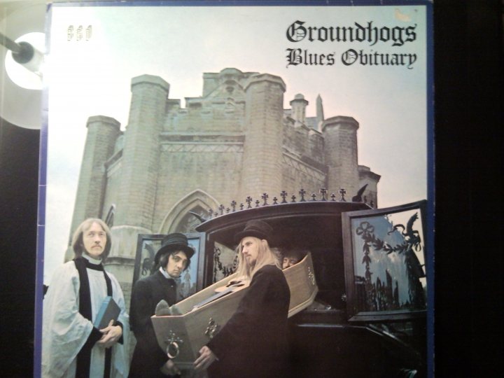 Vinyl Fine Pistonheads - The image is a photograph of a music album cover featuring a group of individuals standing with an open coffin. The central theme of the cover is illustrated by the procession of figures wearing what appear to be clerical garments, reminiscent of gothic architecture, suggesting a religious context. The title "Groundhogs Blues Obituary" is displayed in a gothic-style font, indicate that this is a musical release. The cover has a retro, possibly vintage, aesthetic, with a focus on moody, somber colors.