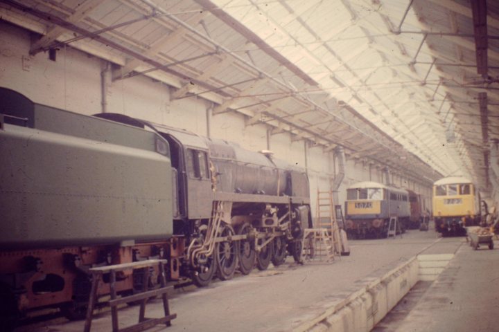 Loco sheds and other railway buildings.... - Page 4 - Boats, Planes & Trains - PistonHeads UK - The image showcases a spacious train depot where several trains are parked. A green, old-style locomotive is prominently displayed in the center of the frame. Surrounding it, there are other trains and possibly a few buses or trucks present. The ceiling appears to be an industrial-grade structure with corrugated metal panels, and natural light is filtering through. This setting suggests a bustling transportation hub.