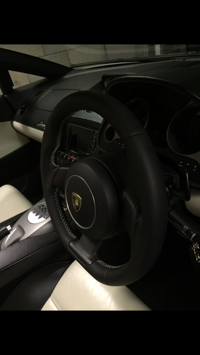 New steering wheel  - Page 1 - Gallardo/Huracan - PistonHeads - The image features the interior of a car, showcasing the driver's side. The primary focus is the black steering wheel with the iconic logo of a prancing horse in the center, indicative of a premium brand like Ferrari or Maserati. The wheel is mounted on a dashboard which also has other controls, likely including radio, air conditioning, and infotainment system. The driver's side appears to be equipped with a seatbelt system visible below the wheel, and the overall color tone of the interior seems to be black or a dark, possibly chrome or titanium, metal surface. The lighting in the picture suggests the car is parked, possibly within a showroom or a garage, with artificial indoor lighting illuminating the interior.