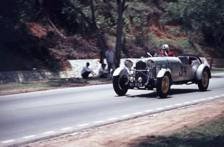 lagonda - Page 1 - Classic Cars and Yesterday's Heroes - PistonHeads - The image captures a scene on a small, rural road. A vintage Bugatti car, its body gleaming against the soft light, is the center of attention, driving down the road with its front light illuminated. A young couple is enjoying a joyride in the car, sharing a moment away from the hustle and bustle of urban life. On the side of the road, a few people in casual attire can be spotted observing the spectacle. The road itself is flanked by a stone wall on one side and a grassy bank on the other, adding to the rustic charm of the scene.