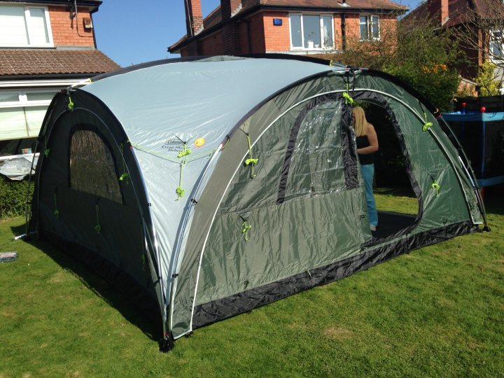 A red car is parked in a field - Pistonheads - The image displays a green and grey tent pitch in a grassy area. The tent is a cabin type model, with a visible base and a movable stretcher creating the arched roof. There are two vents visible on the roof of the tent. One side of the tent has partial mesh of the inner section, and fastening straps are visible on both the roof and base sections, but no handles or pegs at either end. The surrounding grass is a standard green hue, suggesting it might be spring or summer. There are no visible texts or logos on the tent. In the background, there's a view of a residential area, with brick houses, a tree, and a garden visible, indicating an outdoor or backyard setting.