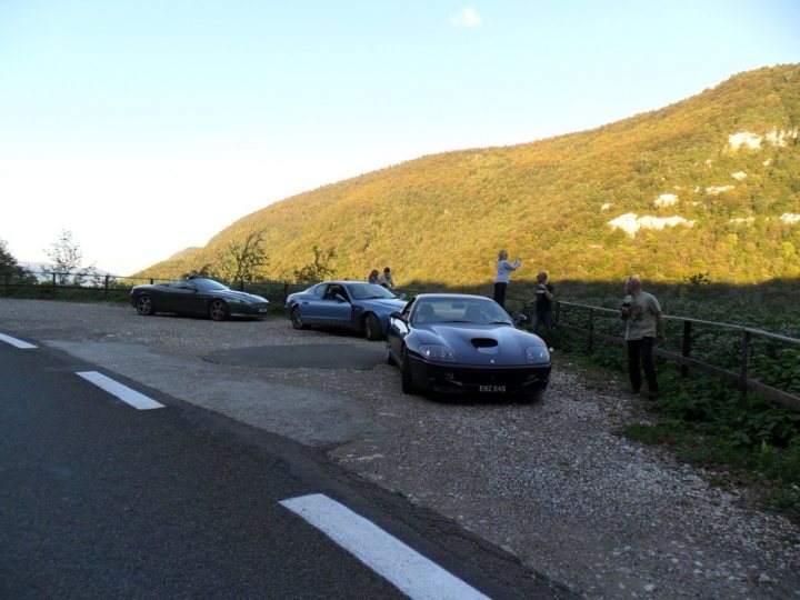 Back from Europe - Page 1 - Aston Martin - PistonHeads - The image captures a serene scene on a winding mountain road. The street is lined with lush greenery, and the open sky above is clear, suggesting a beautiful day. On the side of the road, three individuals are enjoying the view, standing near a fence. They are surrounded by an array of cars, including a striking blue sports car, adding a pop of color to the scene. The location appears to be a popular spot for car enthusiasts, given the numerous cars parked on the side of the road.