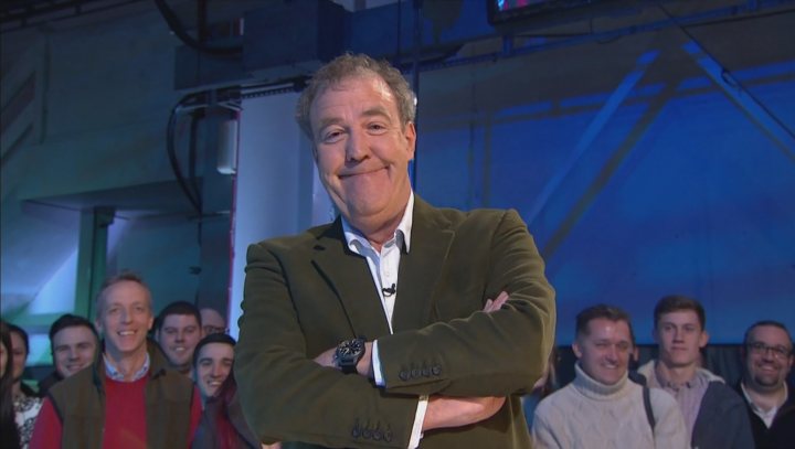 A couple of men standing next to each other - Pistonheads - The image is a still from a television show featuring an older man with greying hair crossing his arms over his chest. He is wearing a dark suit and smiling broadly at the camera. The background is dark, but illuminated by blue light, suggesting a studio setting. It appears to be a promotional image, as there are several people in the backGROUND who seem to be part of a live audience or a show's production crew.