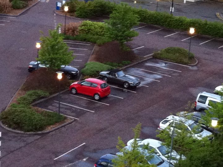 A car driving down a street next to a traffic light - Pistonheads - The image shows a parking lot filled with various cars parked at various angles. The focus of the image is a prominent red car, which is positioned in the center of the parking lot. Surrounding the red car are several other vehicles, some of which appear to be parked closer to the camera. Additionally, there are light posts visible in the parking lot, adding a sense of depth and scale to the image. The area seems quiet with no people visible in the captured frame.