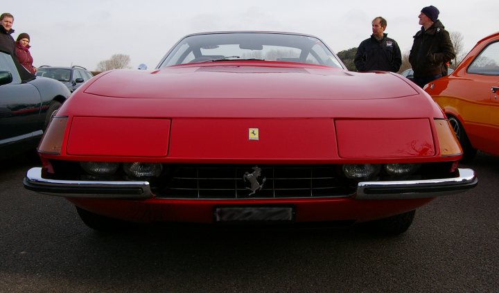 Goodwood Breakfast Club - Sushi Sunday - photos - Page 1 - Goodwood Events - PistonHeads - The image showcases a red Ferrari car parked in the middle, surrounded by several other vintage cars. The Ferrari features a license plate at the front of the vehicle. There are three people standing around the cars, possibly admiring the design and craftsmanship. The setting appears to be an outdoor event or a similar gathering where vintage cars are on display. The colors, shapes, and design details of the Ferrari and the surrounding vintage cars indicate a time when such vehicles were popular and luxury cars.