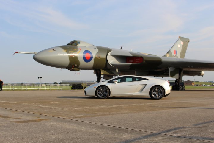 Pistonheads - This image captures an interesting juxtaposition of old and new technology. There's a military aircraft with a camouflage pattern and distinctive roundels, suggesting it could be a fighter jet. Parked beside it is a sleek white sports car, which contrasts with the military aesthetics of the jet. Both the aircraft and the car are quite large, occupying a significant portion of the image, and appear to be in a landing strip area. There's a single person in the vicinity, giving a sense of scale to the scene. The background is partly cloudy, providing depth to the image. The setting suggests a possible air show or exhibition, where military and personal vehicles are displayed together.