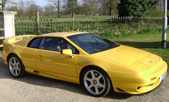 Esprits needed for Hedingham - Page 1 - Esprit - PistonHeads - The image captures a vibrant yellow sports car parked on a gravel area by a fence. The car, which is the main subject of the image, is facing towards the left side of the photo, placed on a patch of grass. The background features a lush green field, with trees dotting the landscape. The presence of a fence suggests a boundary or protection for the area where the car is parked. The image is taken during the day, as indicated by the visible profile of the sun.