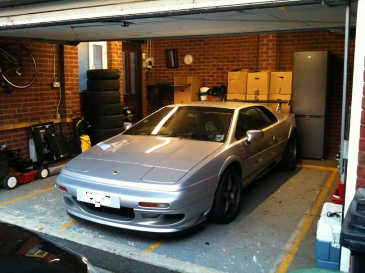 Single Garage Pistonheads Fit Esprit - The image shows an indoor garage where a police car is parked. The car is metallic and has distinctive markings and text on it, although the license plate is hanging on hood. To the right of the car is a clock mounted on the brick wall, which adds a sense of time and routine to the scene. In the background, one can find a collection of various household items, perhaps indicating the garage's multi-purpose use. The environment suggests an everyday life setting with the garage likely being part of a residential home.