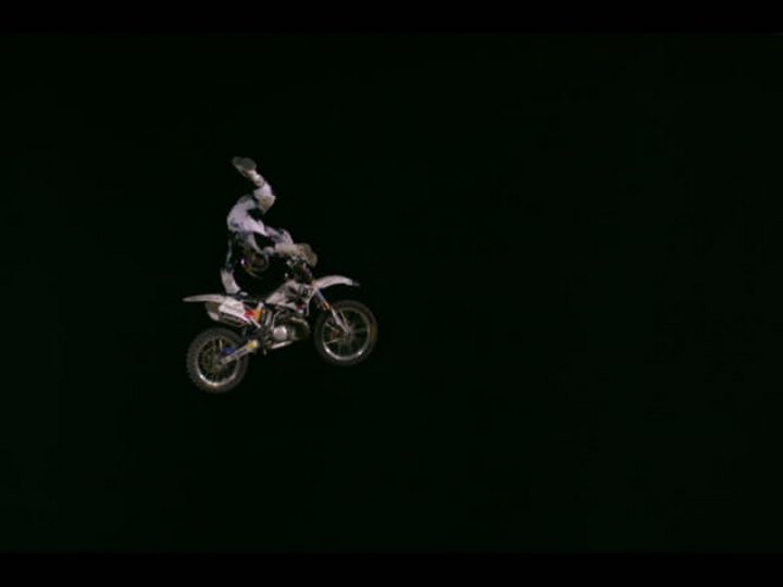 Pistonheads Fighters Bull Red - The image depicts a daring motorcycle stunt. A motorcyclist, clad in full riding gear, is captured in mid-air while performing a wheelie trick at night. The rider and motorcycle are the main focus of the image, standing out against the stark black background. The motorcycle is in motion, hovering above the ground, and the rider's lean and grip on the handlebars suggest skill and control. The dark surroundings emphasize the daring nature of the stunt.