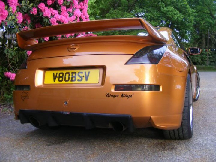 A red car with a surfboard on top of it - Pistonheads - The image presents a scene with an orange car parked on a street. The car is not significantly visible due to its private license plate, which is only partially visible. The car's license plate is yellow and displays letters and numbers, but the registration details are not visible to maintain privacy. There is a pink flowering bush visible in the background and another car parked further down the street. There is a small amount of text written on the car, but due to the angle and focus of the photo, it's difficult to read precisely what it says. The surroundings include a walking path parallel to the road, suggesting this could be a residential or recreational area. The road appears to be in good condition with visible white lines, indicating a designated road for vehicles.