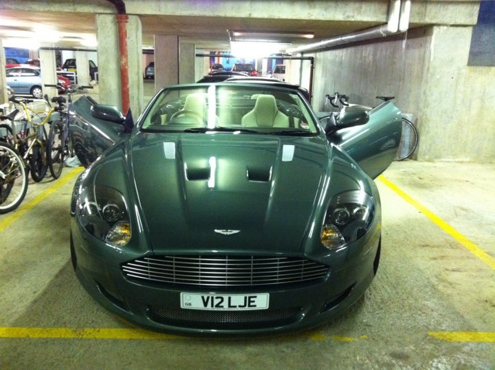 Pistonheads - The image shows a sleek, dark green Aston Martin car parked under a multi-level parking structure. The car's boulevard design suggests it's a sports or luxury sedan. In the background, there's a bicycle stand with multiple bikes. The concrete floor of the parking structure is marked with yellow lines, indicating spaces and pathways. The lighting is dim, giving the scene a subdued atmosphere.