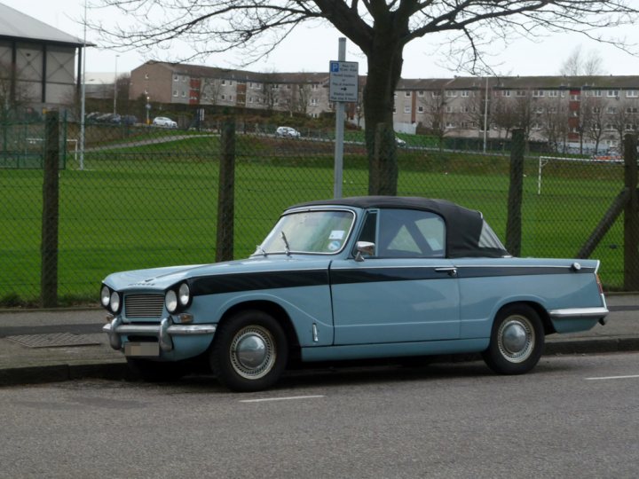 Pistonheads - In the image, a black and blue convertible car is parked on the side of a road. The car is positioned parallel to the curb, indicating a legal parking spot. Beyond the road, a green field stretches out, dotted with cars in the distance, suggesting a populated area or a parking lot associated with the road. A silver four-door sedan can also be seen further down the road, implying recent or ongoing traffic. The scene is simple yet colorful, with the vibrant blue car as the central focus.