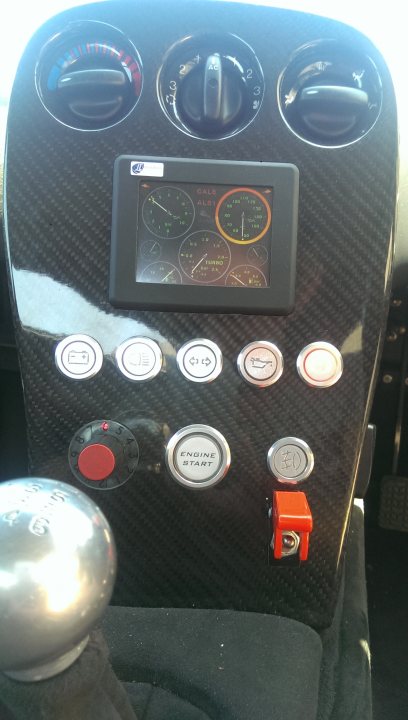 The padded thing in front of the passenger - Page 1 - Noble - PistonHeads - The image portrays an intricate automotive dashboard. The focal points are the multitude of screens and knobs that are uniquely arranged. The predominant color scheme is black with white and red features, possibly indicative of digital readouts and buttons for function controls. The screens display a variety of icons, likely serving different purposes, while the knobs are positioned beneath them for easy access. The overall layout suggests a luxurious and technologically advanced vehicle interior.