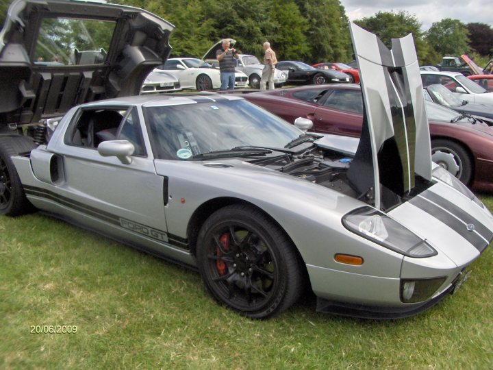 Pistonheads Raleigh Rally - The image showcases a car show with a close-up view of a silver convertible parked on grass. The convertible is the centerpiece of the image, with its hood open revealing the engine. People are standing behind the car, and other cars with open hoods can be seen in the background. The setting appears to be an outdoor event.