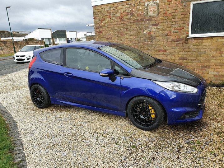 Fiesta ST MK7.5 Track car  - Page 31 - Readers' Cars - PistonHeads UK - The image shows a blue Ford Fiesta parked on a driveway. It appears to be a new model car with a black roof and a white stripe along the side, which is a common customization for Fiestas. The vehicle has a set of distinctive rims and blacked-out windows. In the background, there's a building that seems to be residential or possibly a small business establishment. The overall setting suggests a quiet suburban neighborhood.