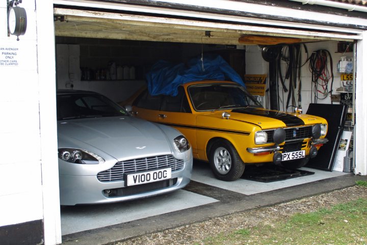 Our other cars - Page 10 - Aston Martin - PistonHeads - The image depicts a garage with two vintage cars. On the left side of the image, we see the rear of a metallic silver car with a distinctive grille and "VIOODOG" text. To the right is a vibrant yellow and black striped car with the number "PX 551" on it. The garage appears well-maintained, with tools and products hanging neatly on the wall. A notable feature is a large blue tarp, folded and secured over the yellow car, possibly for protection or a planned modification project. These cars seem to be cherished acquisitions, as indicated by their clean, elegant appearance.