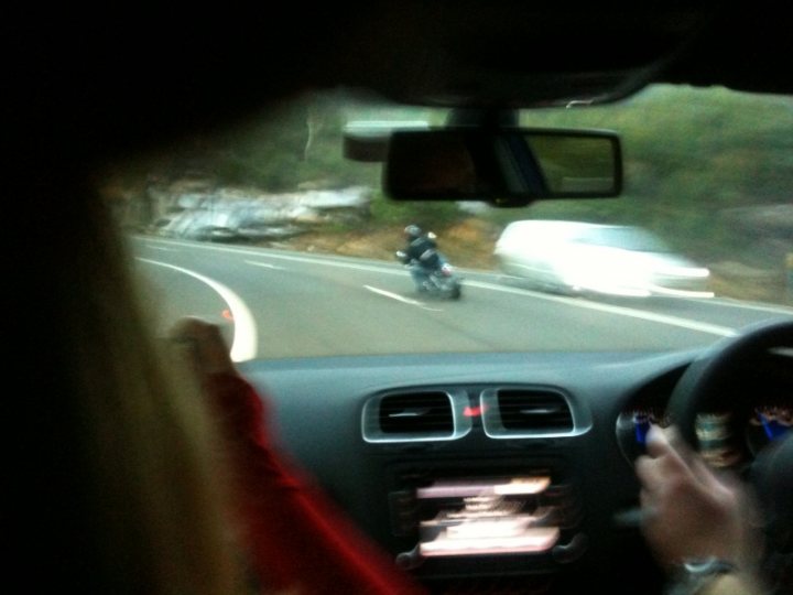 Pistonheads - The image depicts a motorcycle driver travel-viewed through a car windshield. The scene suggests a dual carriageway with a van on the far right and a vehicle on the opposite carriageway. The speedometer on the car's dashboard displays a red needle, indicating a high speed, and the person driving the car appears to be using the steering wheel on the right side.