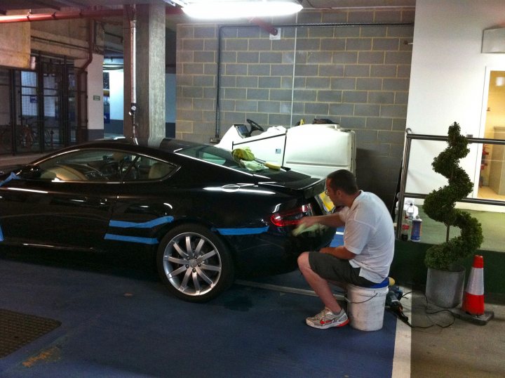 1 day detail - Page 1 - Aston Martin - PistonHeads - In the image, a man is kneeling down next to a black sports car in an underground parking garage. The car has a neat, dark color and shiny chrome accents, giving it a sleek appearance. The man, with his back to the camera, is wearing a white t-shirt and denim shorts and appears to be polishing the car. In the background, there's a potted plant and a garage door, adding to the urban setting. The floors of the garage are a solid slab of concrete, and it looks like a quiet, secluded parking spot.